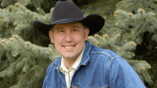 Smiling person in a black cowboy hat and blue jacket in front of evergreen trees