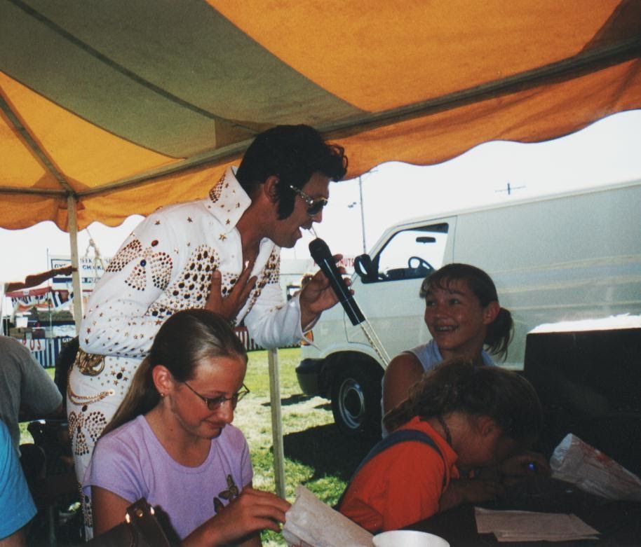 Person speaking into a microphone at a table under a yellow canopy, with others seated nearby outdoors