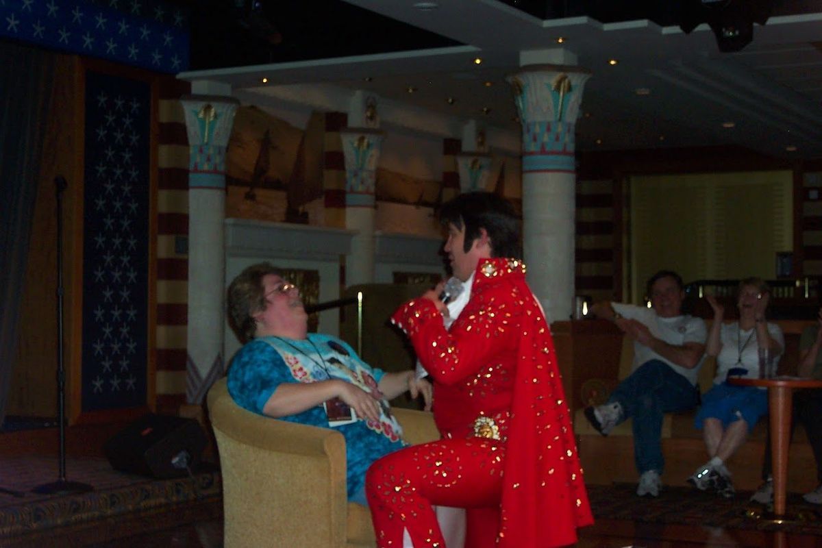 Elvis impersonator in red suit serenades a seated woman in a dim restaurant.