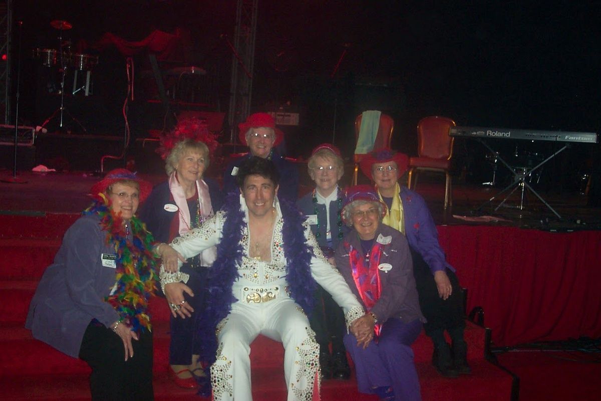 Group of five people posing onstage, wearing bright costumes and leis under red stage lighting.