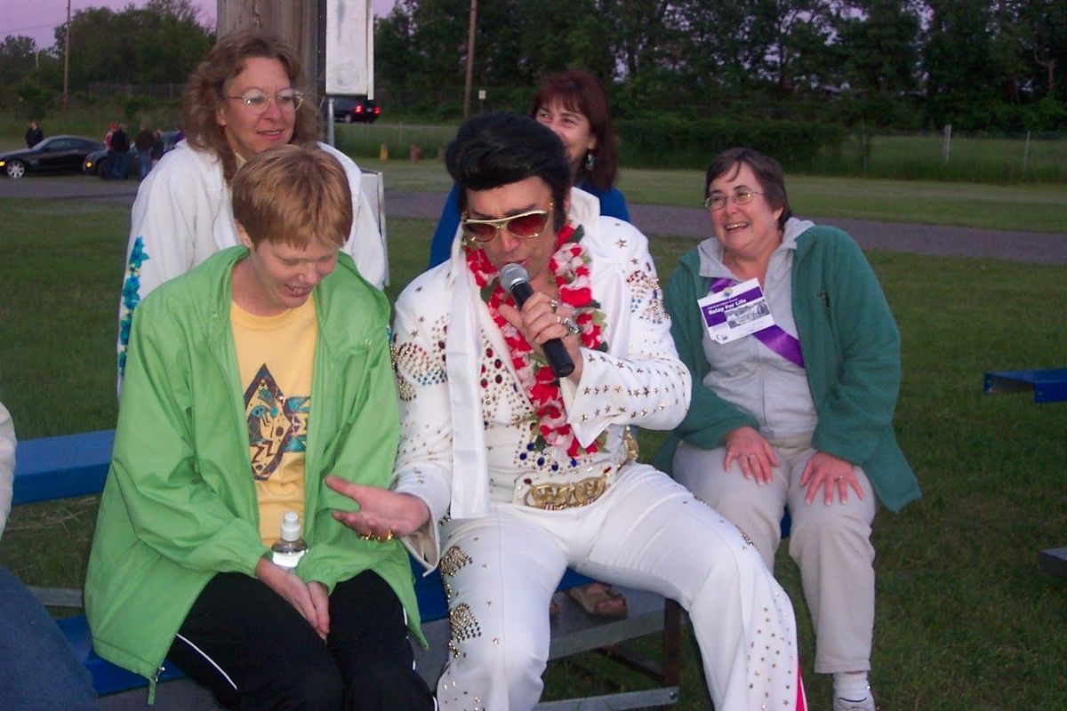 People sitting on a bench outdoors, one in a white Elvis-style outfit, with flowers and a tree in the background