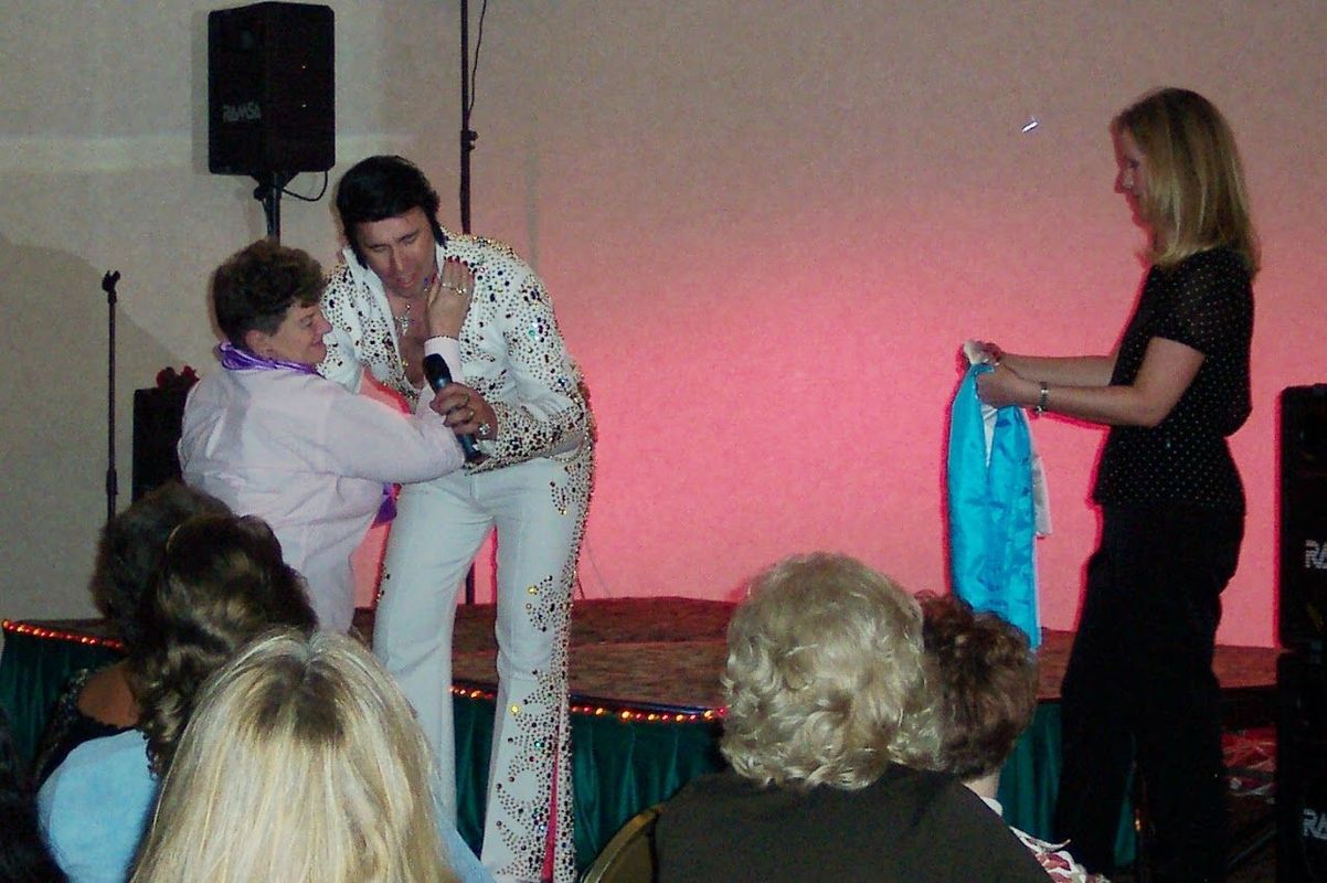Elvis impersonator on stage at a party, receiving a blue gift bag from a guest.