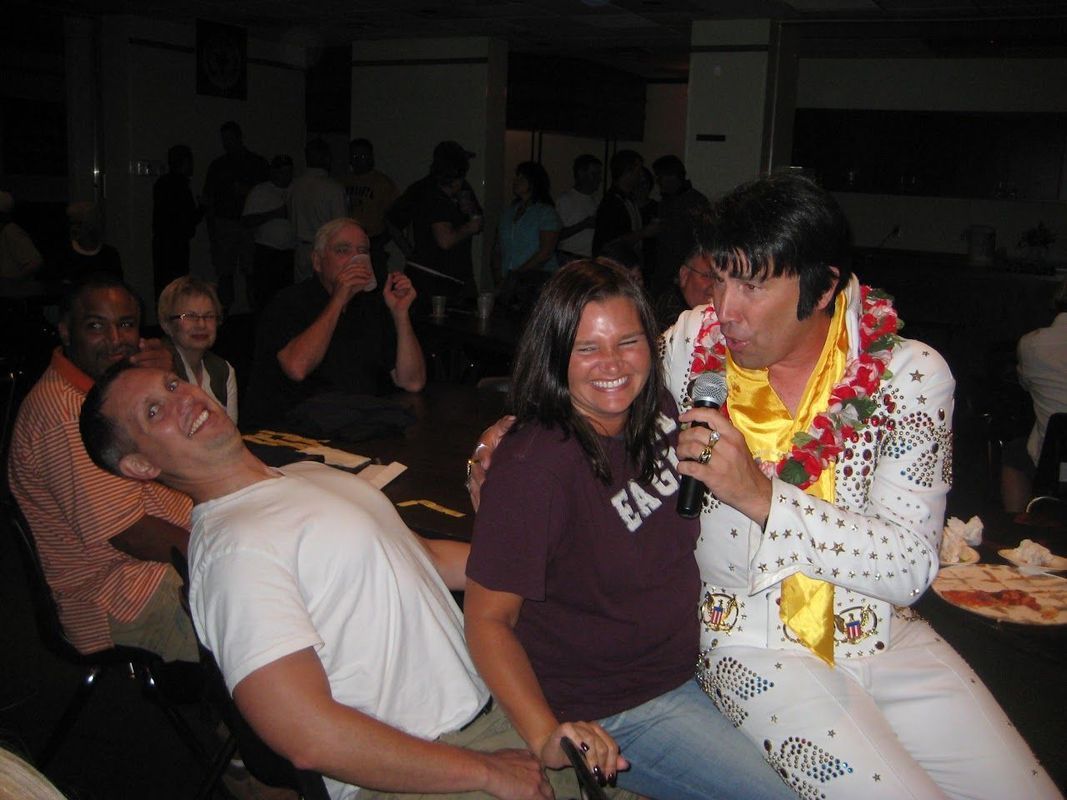 Elvis impersonator performing with smiling audience members in a dim indoor venue