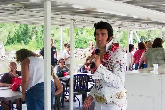 Man in white patterned shirt holding a drink on a crowded outdoor patio with seated people