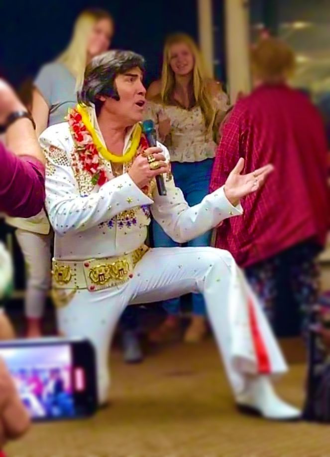 Elvis tribute performer in white jumpsuit singing onstage with microphone and red scarf