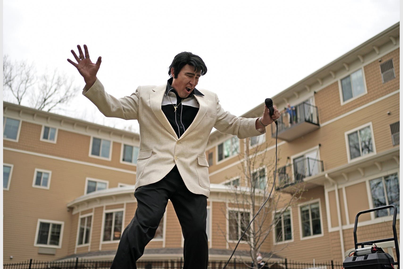 Man in a white jacket holding a microphone, standing in a courtyard with apartment buildings behind him.
