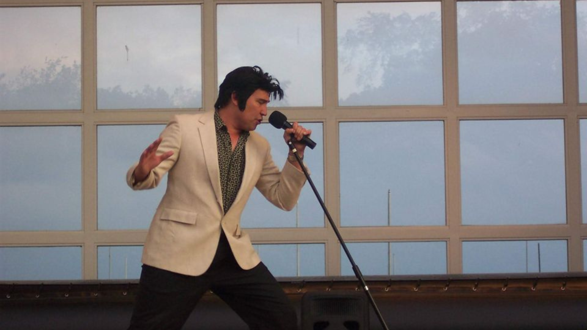 Singer in a cream blazer performing into a microphone on stage with large window panels behind him