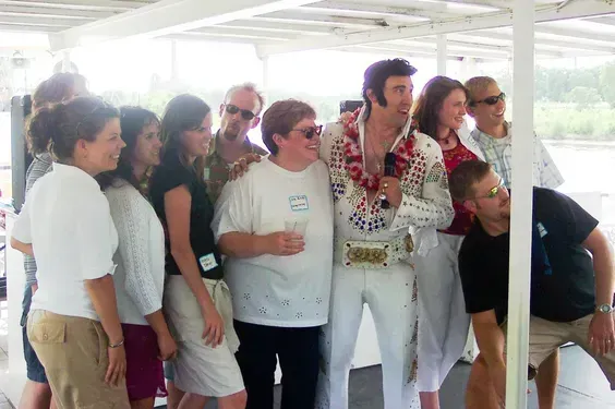 Group of people standing close together at a bright outdoor event, with one man in a white jumpsuit holding a drink.