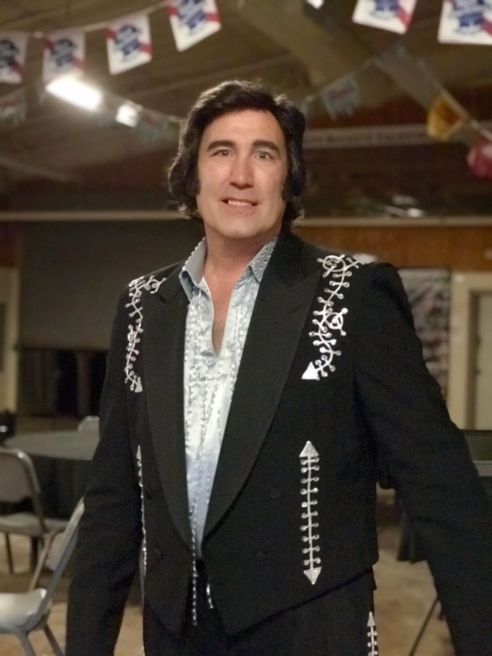 Man in a black embroidered jacket and silver shirt standing indoors, smiling at an event.