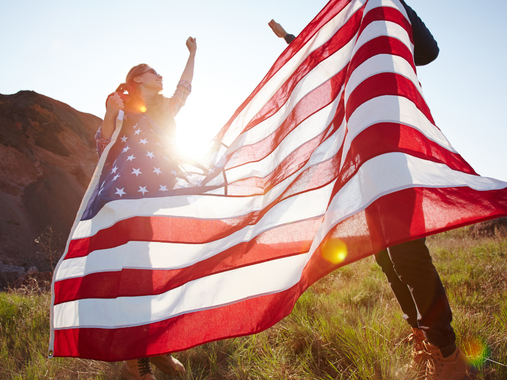 People waving a large American flag outdoors at sunset in a grassy field.