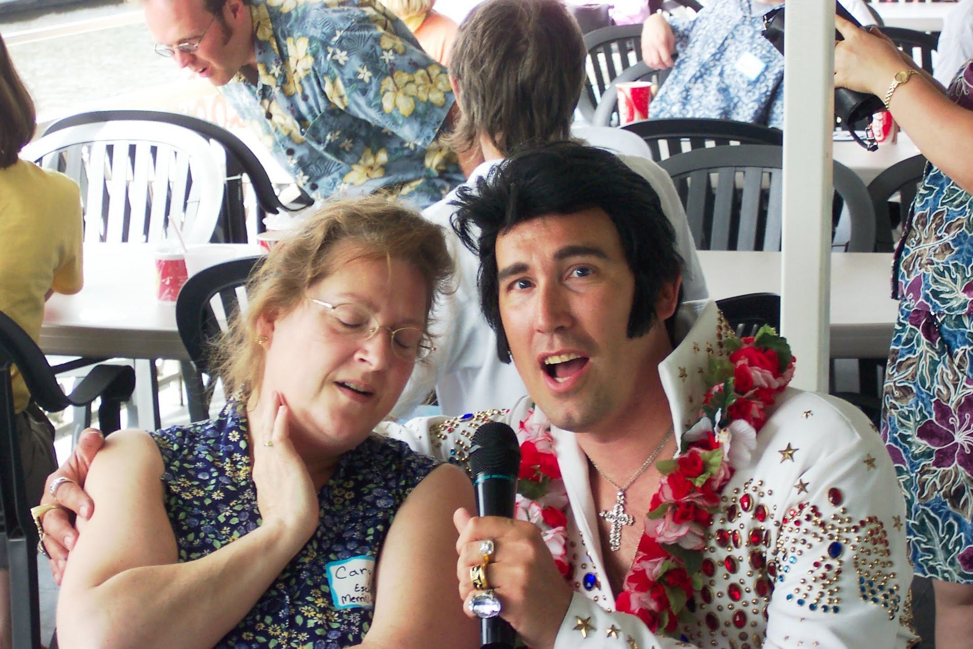 Two people singing into a microphone at an outdoor gathering, wearing leis and seated among white plastic chairs.