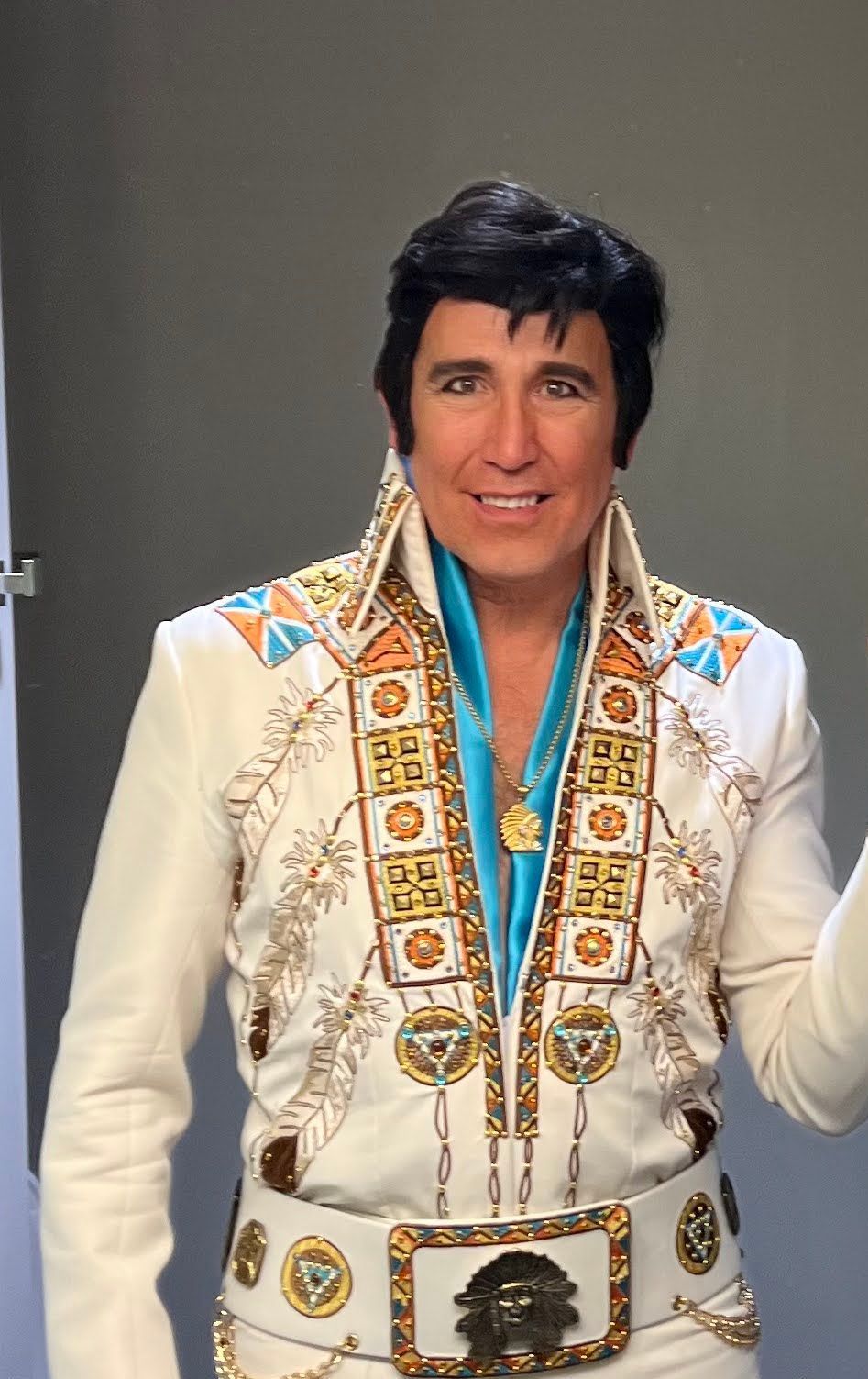 Man in white Elvis-style jumpsuit with turquoise trim and scarf, standing against a gray background.