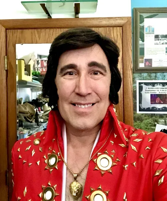 Person in a red jeweled jacket and black wig smiling indoors, with framed items and shelves behind them