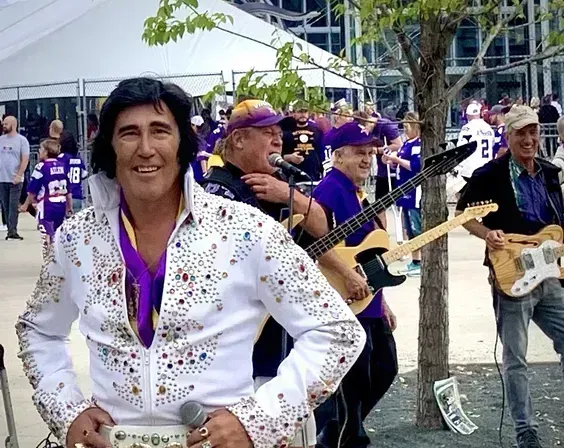 A man in a white sequined Elvis-style suit stands in front of a purple-clad band outdoors.