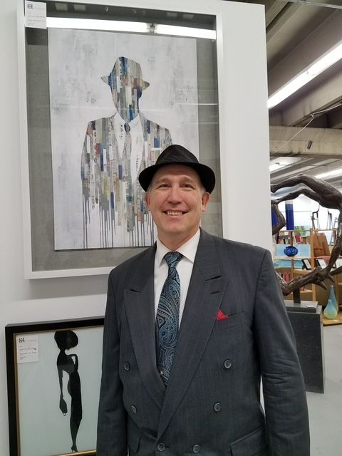 Man in a dark suit and hat standing beside framed artwork in a gallery.