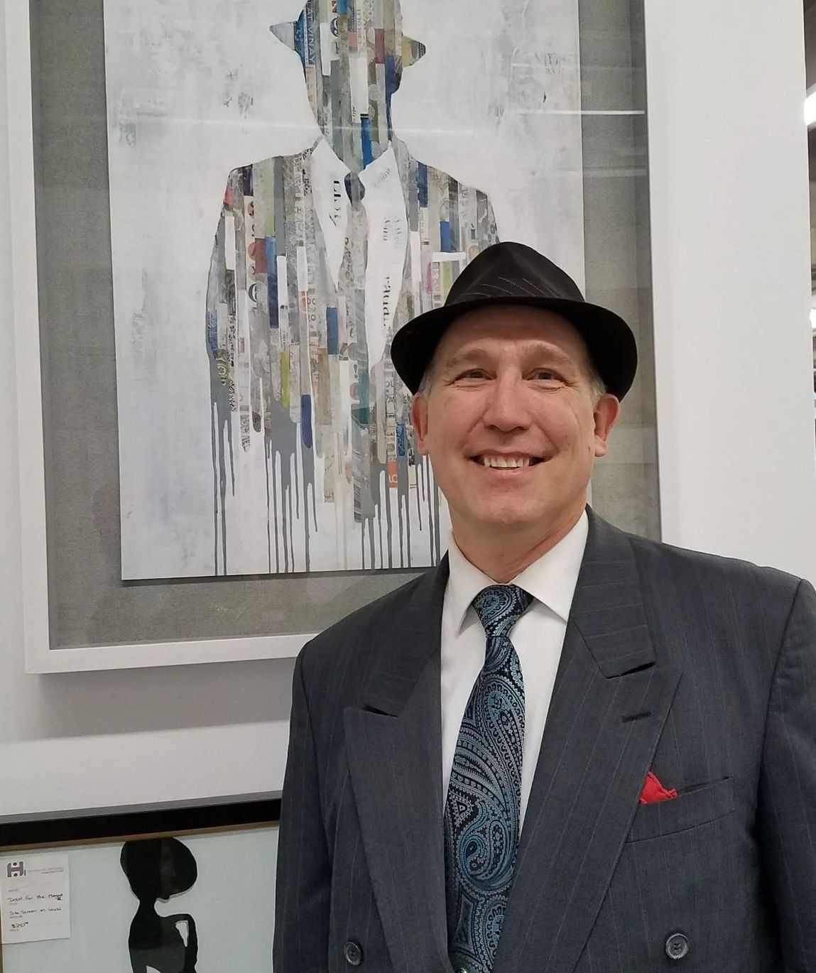 Man in a dark suit and black hat smiling beside framed abstract artwork in a gallery.
