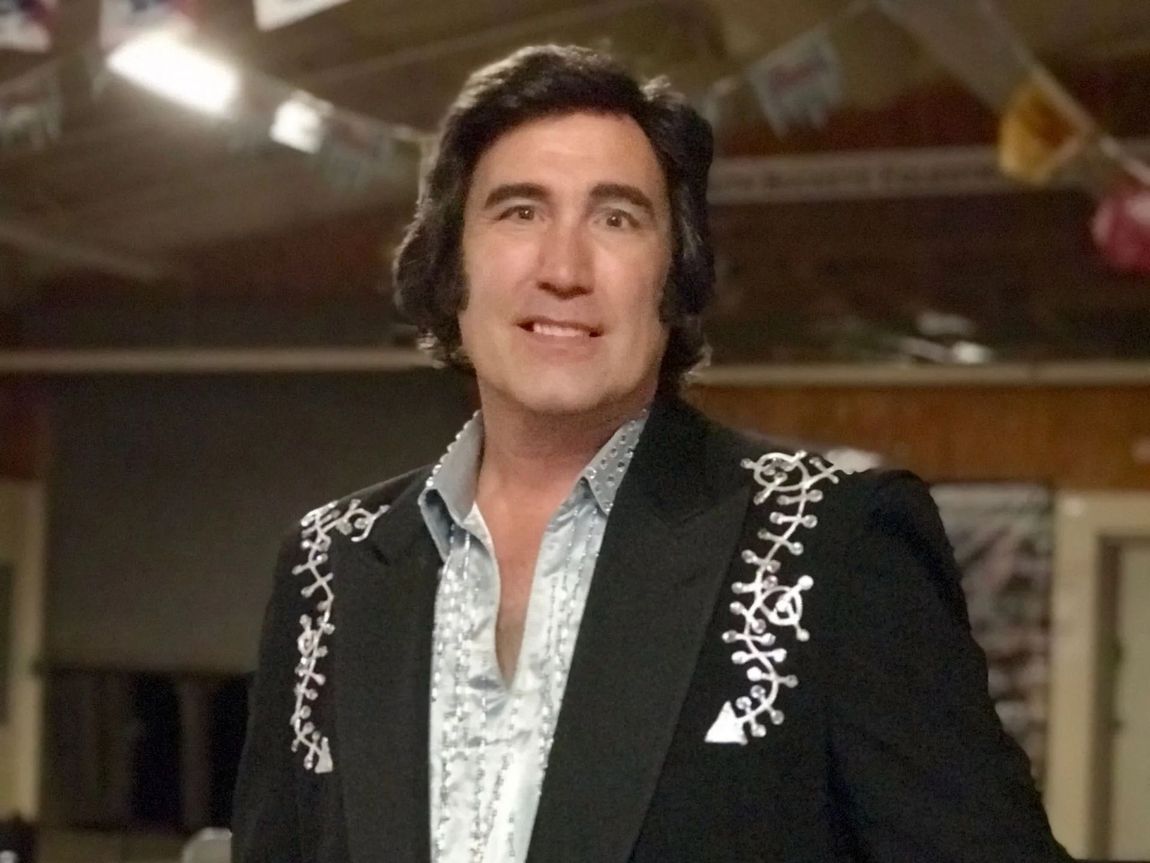 Man in black embroidered suit smiles indoors, with festive decorations in the background
