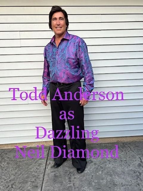 Todd Anderson posing in a purple patterned shirt and black pants against a white siding wall.