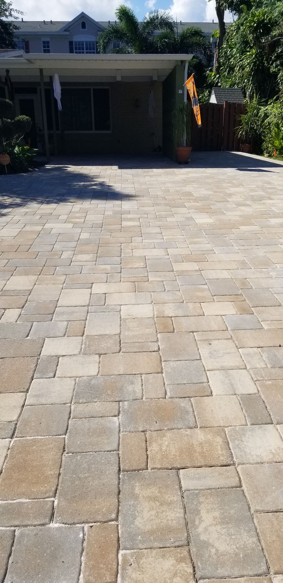 Brick-paved driveway leading to a house with a shaded carport and trees in the background
