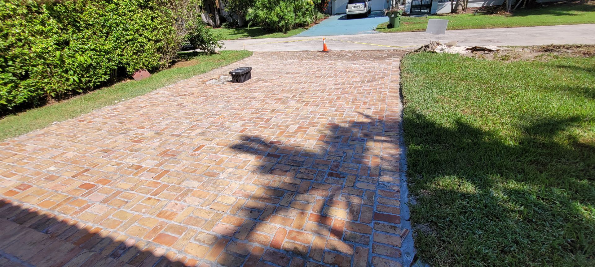 Brick driveway with a long tree shadow, green lawn, and a distant orange cone near the street.