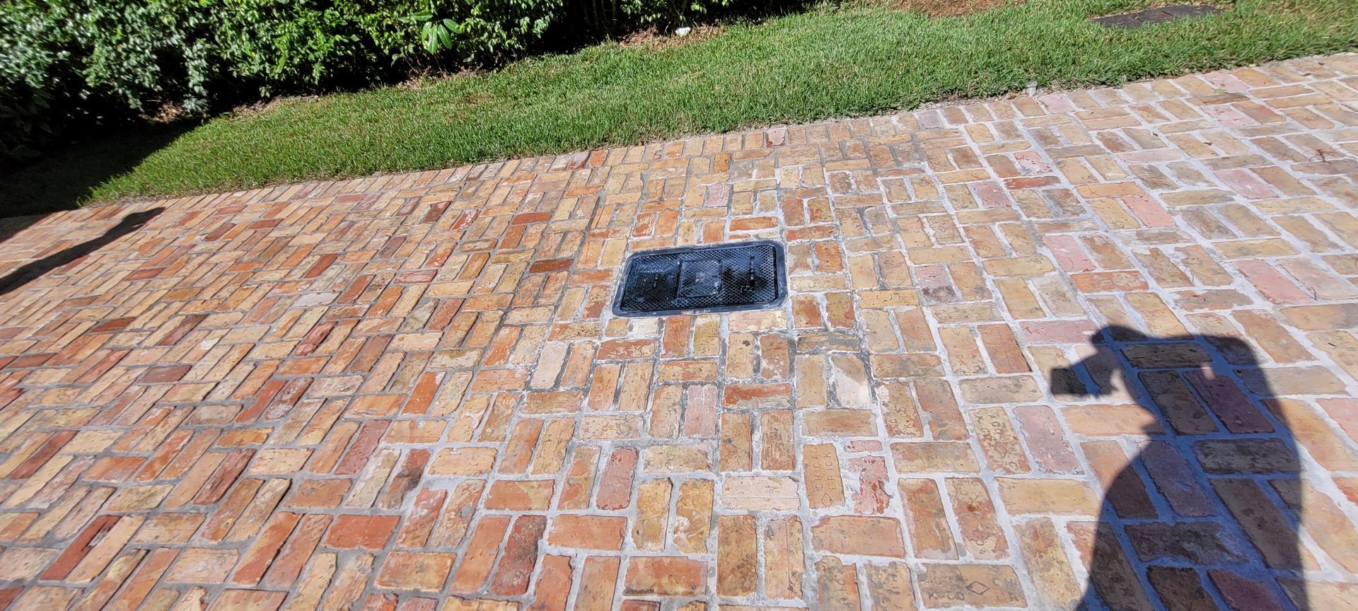Brick patio with a rectangular ground drain and a person’s shadow at the right edge.
