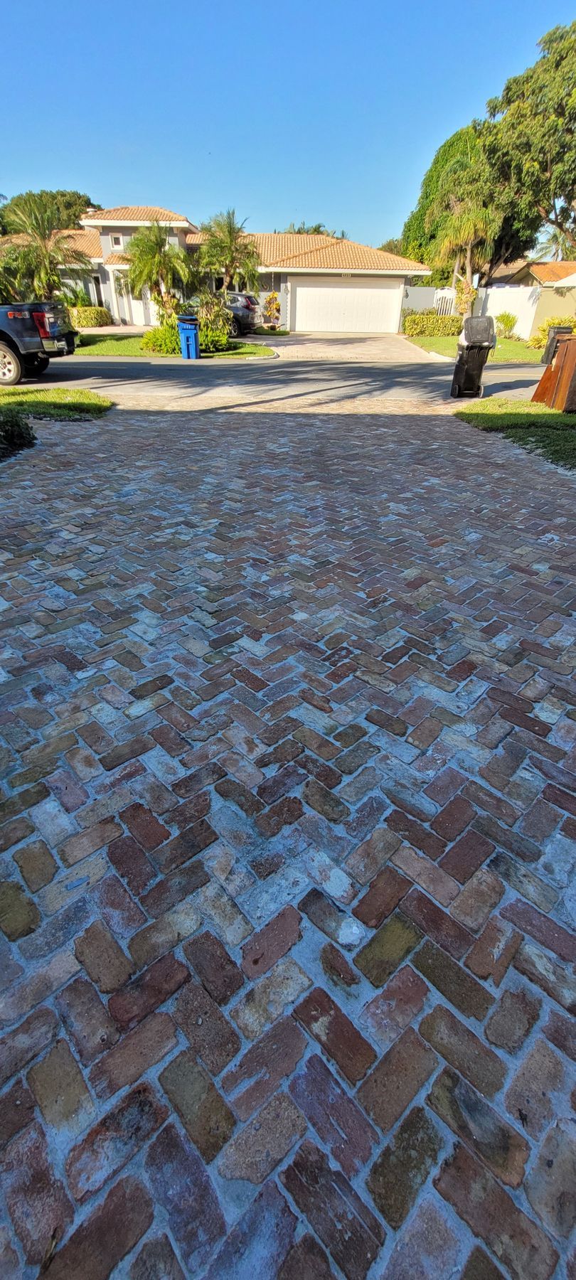 Brick driveway leading to a suburban house with trees and parked cars on a sunny day
