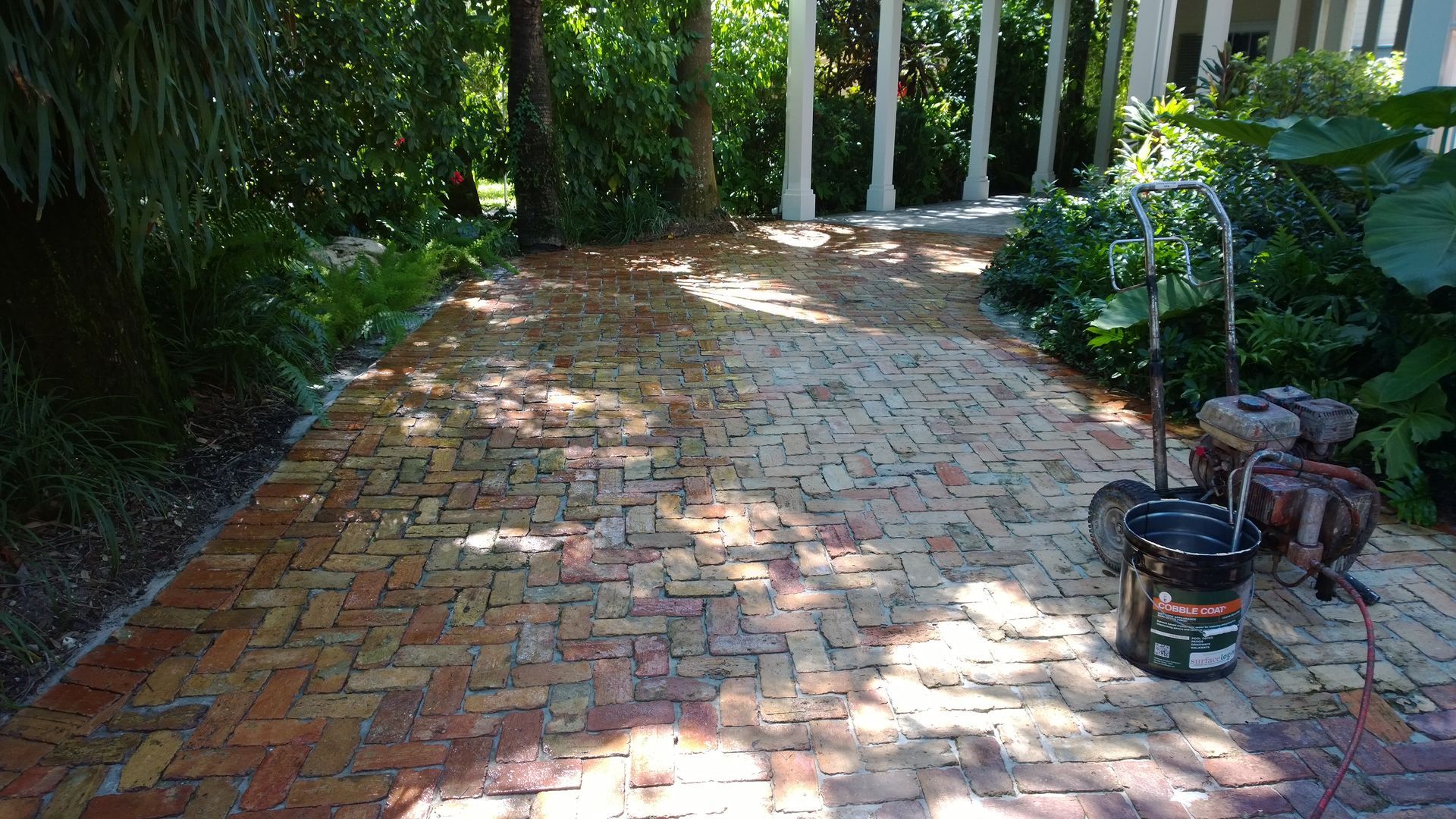 Brick patio driveway with dappled sunlight, bordered by greenery and a fence, with a small tool cart on the right