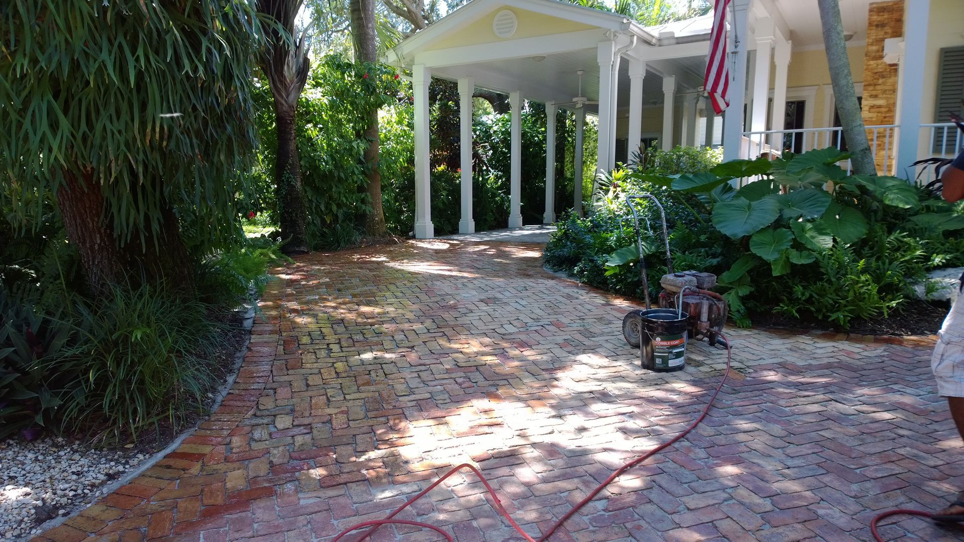 Brick driveway leading to a white columned porch, bordered by lush green plants and trees.