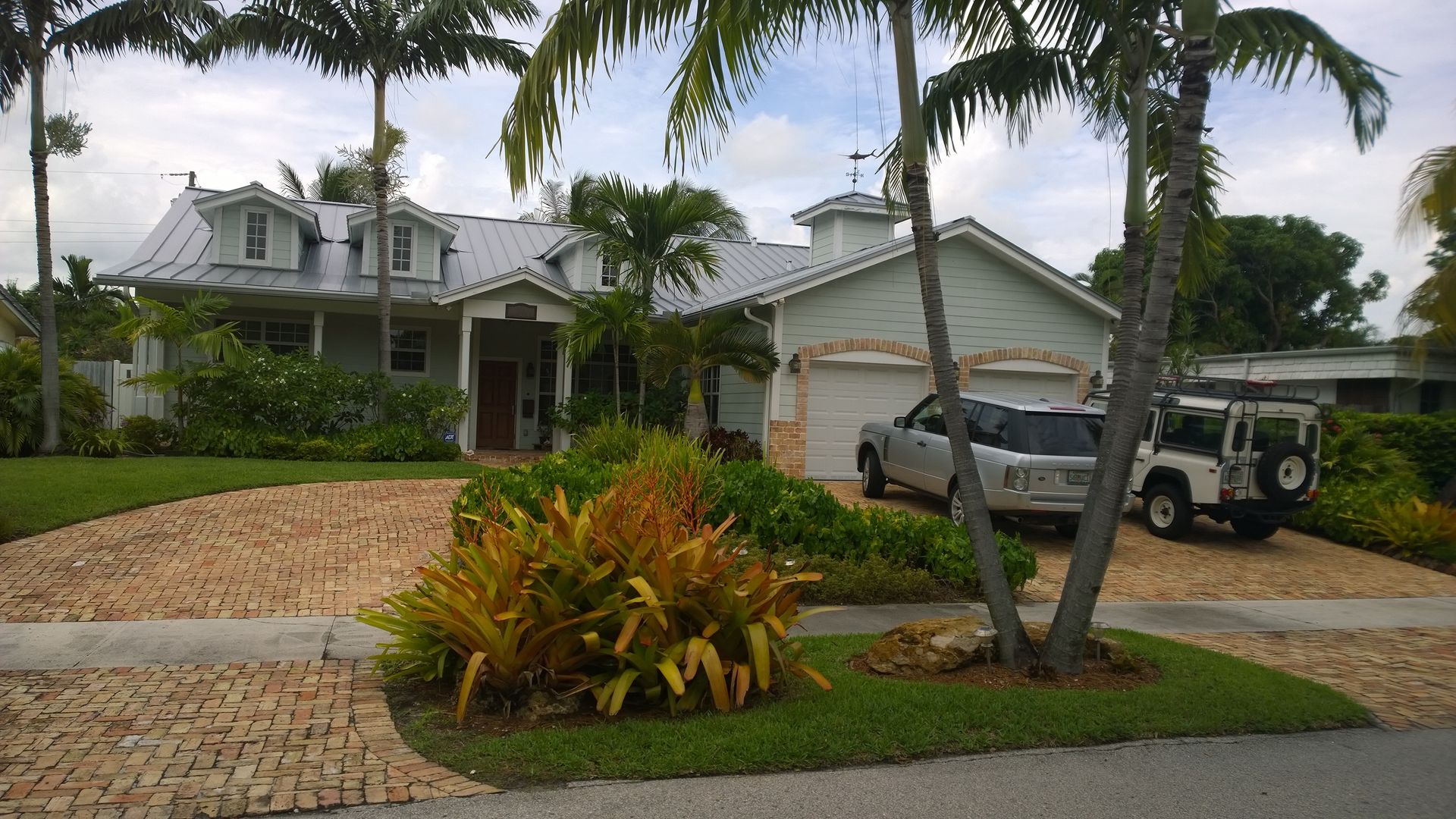 Suburban house with palm trees, a brick driveway, and two parked vehicles under a cloudy sky
