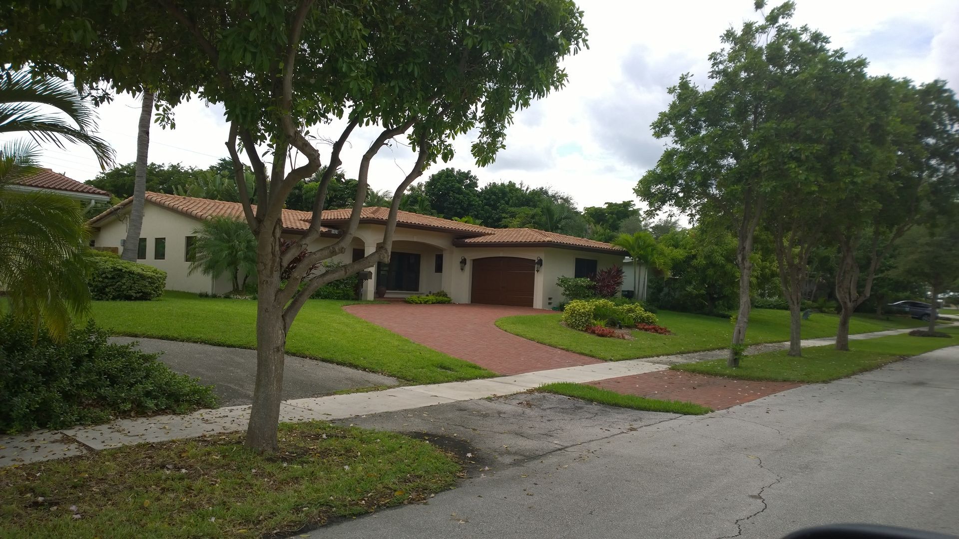 Suburban house with a red-tile roof, brick driveway, and trees along a quiet street
