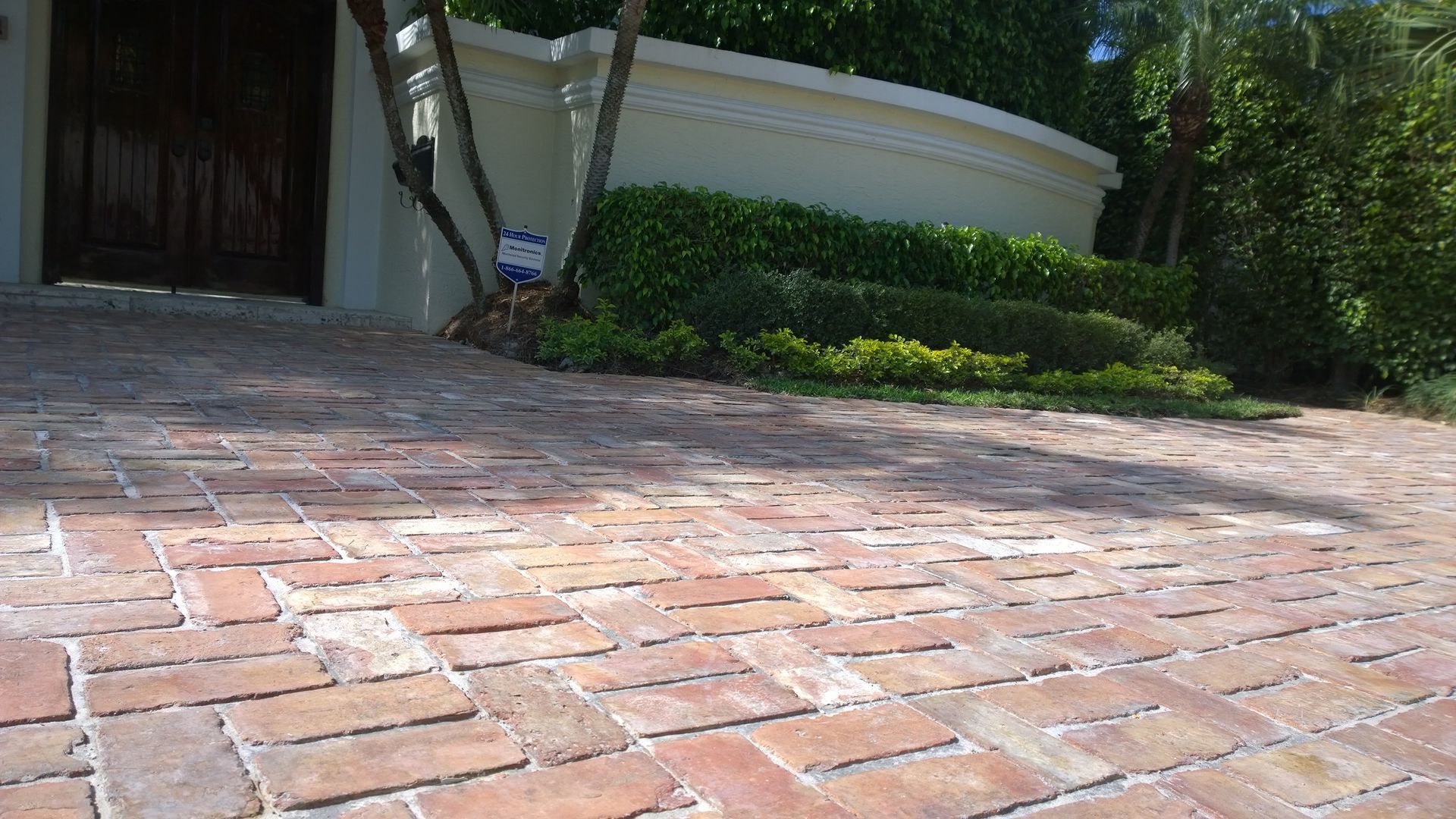 Brick walkway leading to a house entrance with green shrubs and trees in sunlight