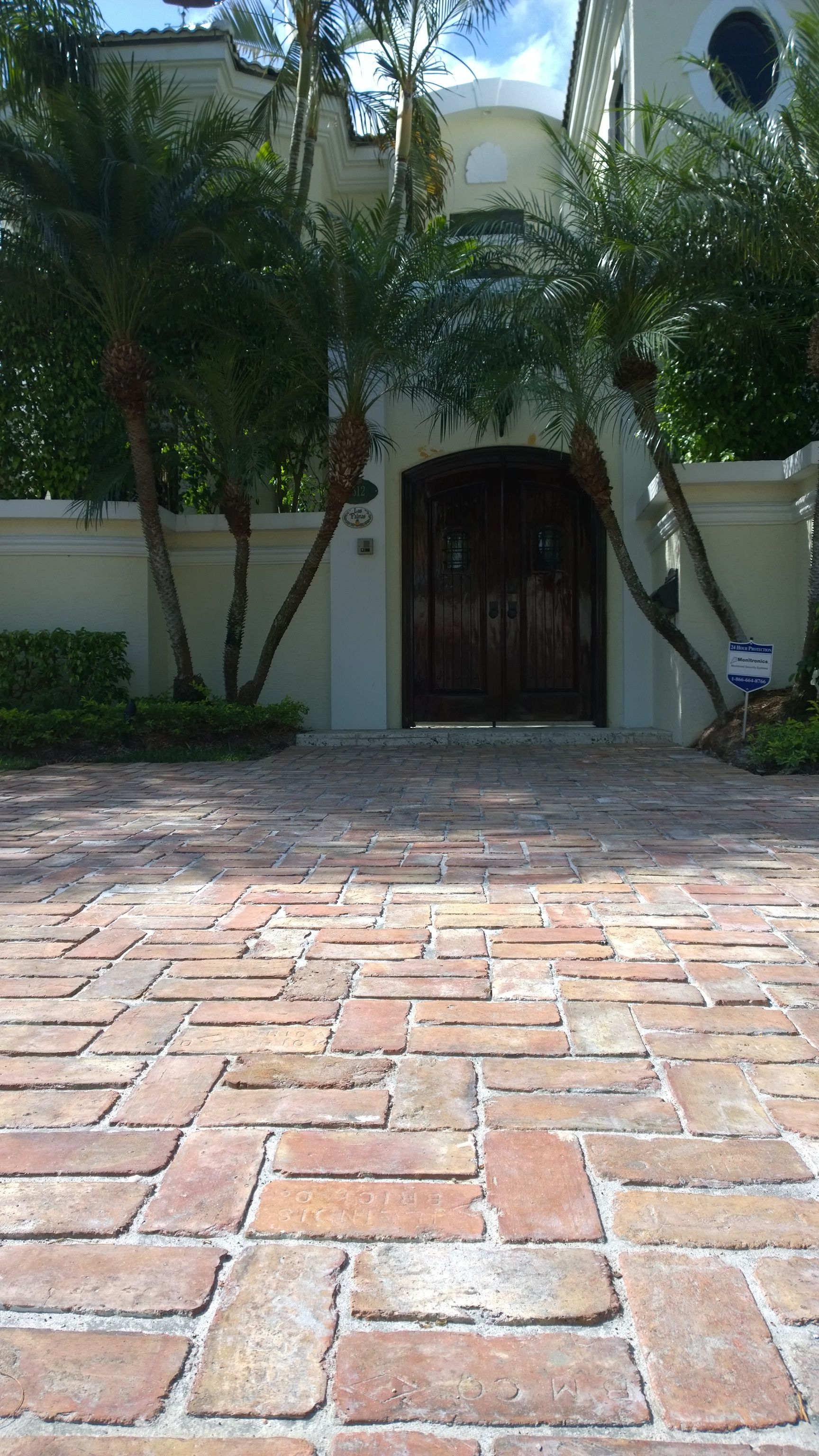 Brick driveway leading to a gated arched entrance with palm trees and white stucco walls