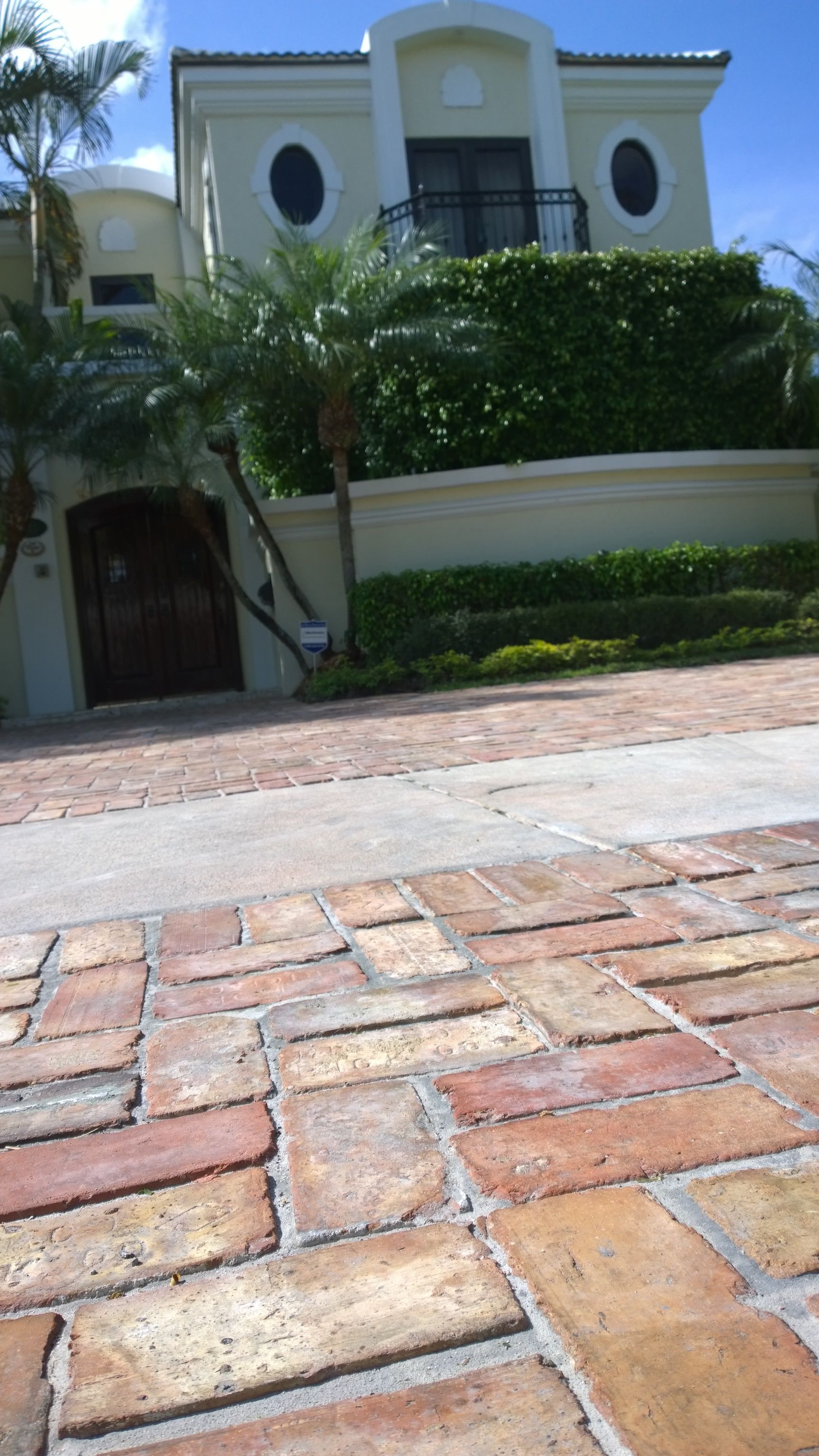 Brick driveway leading to a cream-colored house with palm trees and hedges in front.