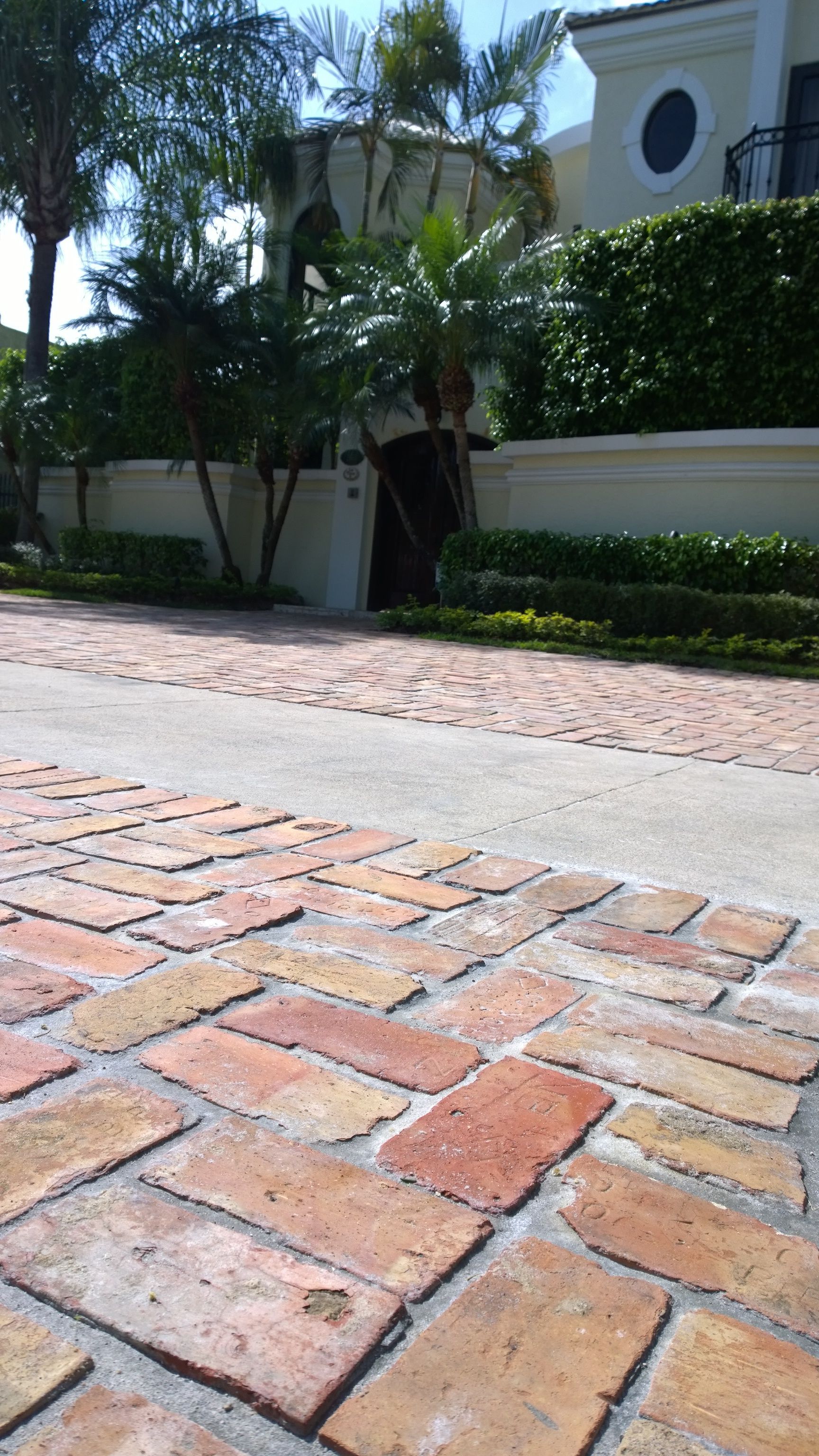Low-angle view of a brick driveway and sidewalk beside a white stucco house with trees and hedges