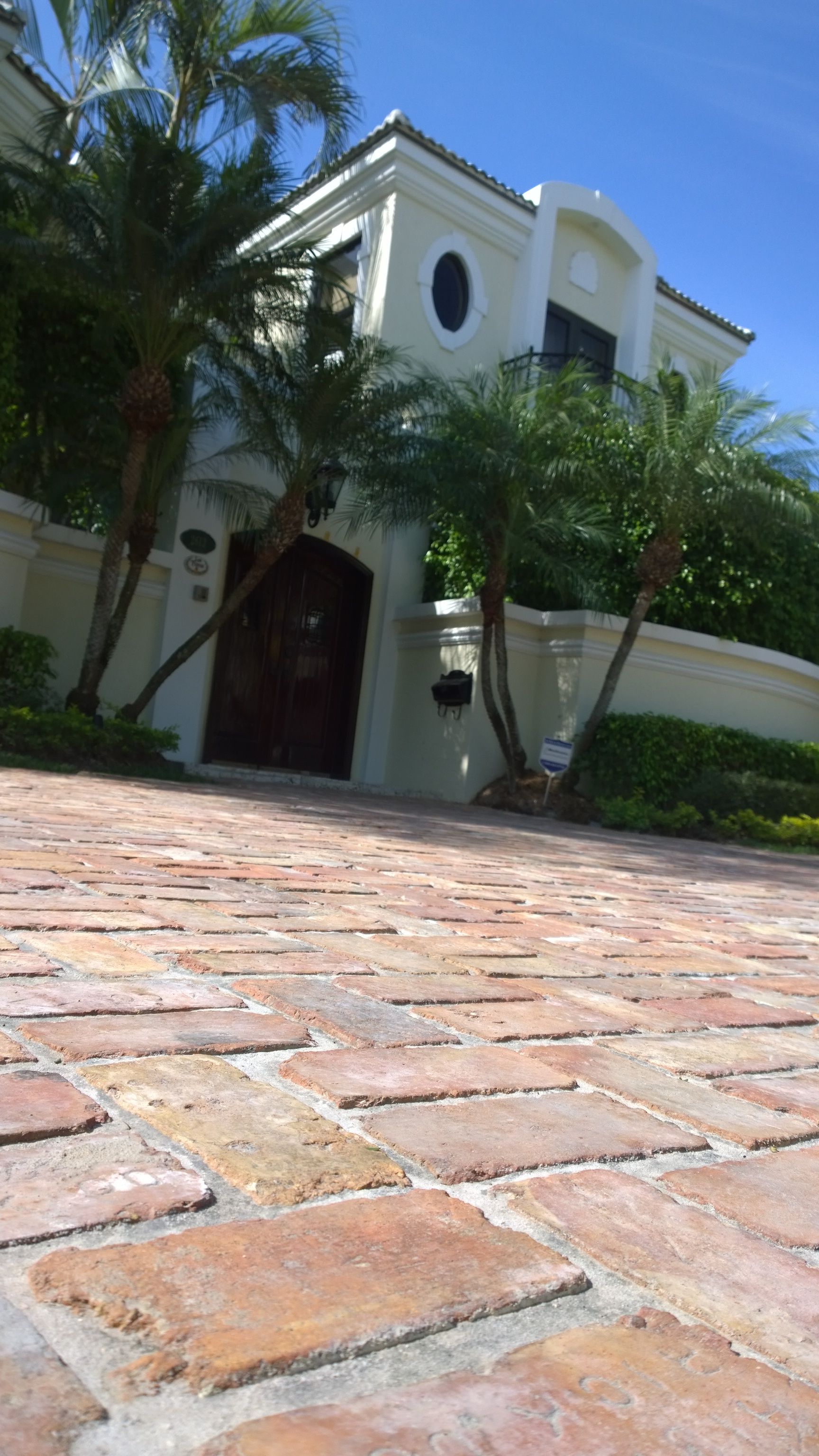 Low-angle view of a white stucco house with arched entry, palm trees, and a brick driveway under a blue sky