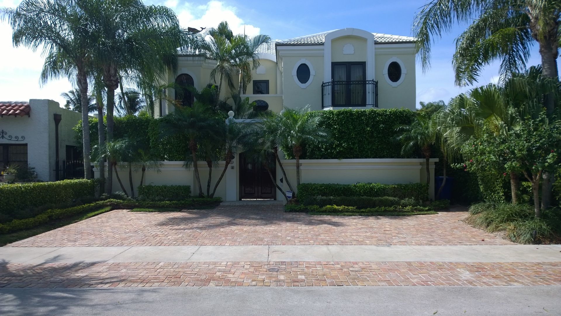 Modern white house with balconies, palm trees, and a paved driveway behind a hedge.