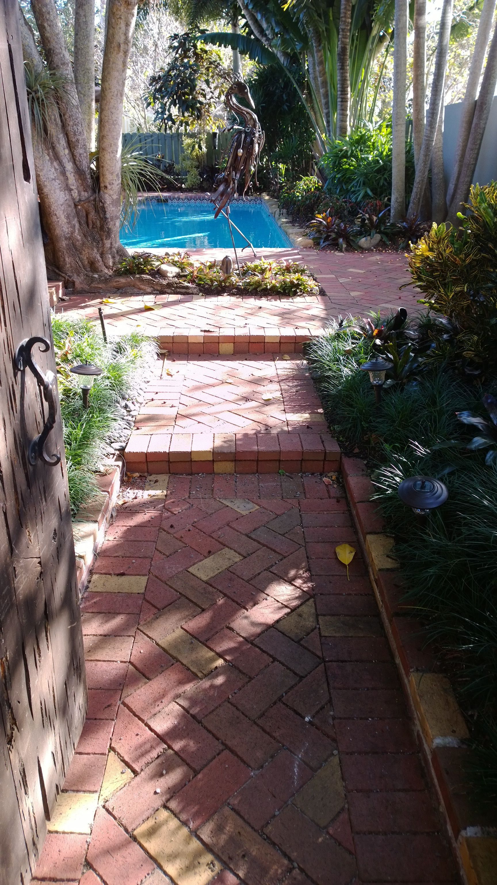 Brick path through a garden leading to a blue pool, framed by trees and shrubs.