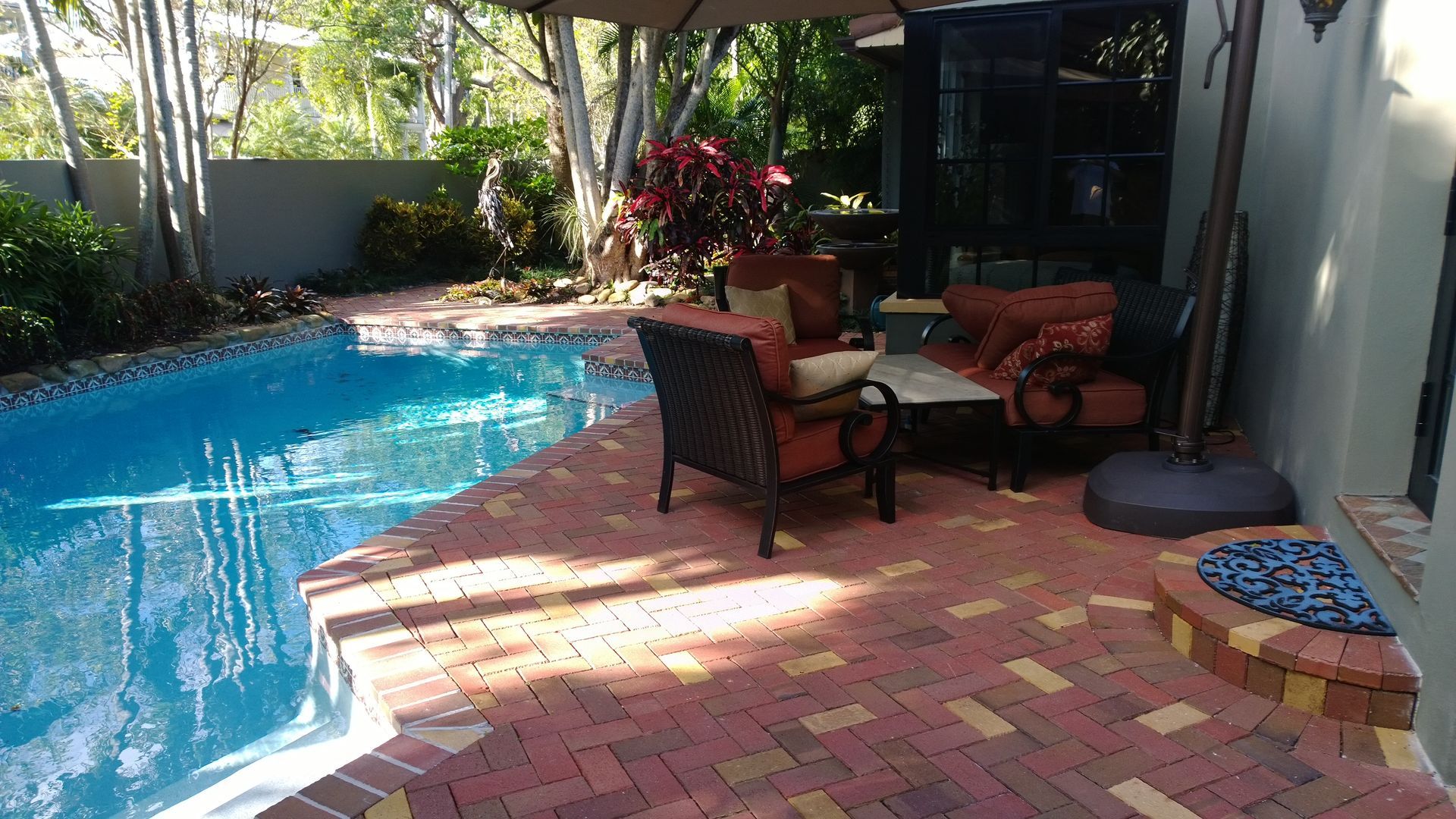 Backyard pool patio with red brick flooring, wicker chairs, and potted plants in bright sunlight
