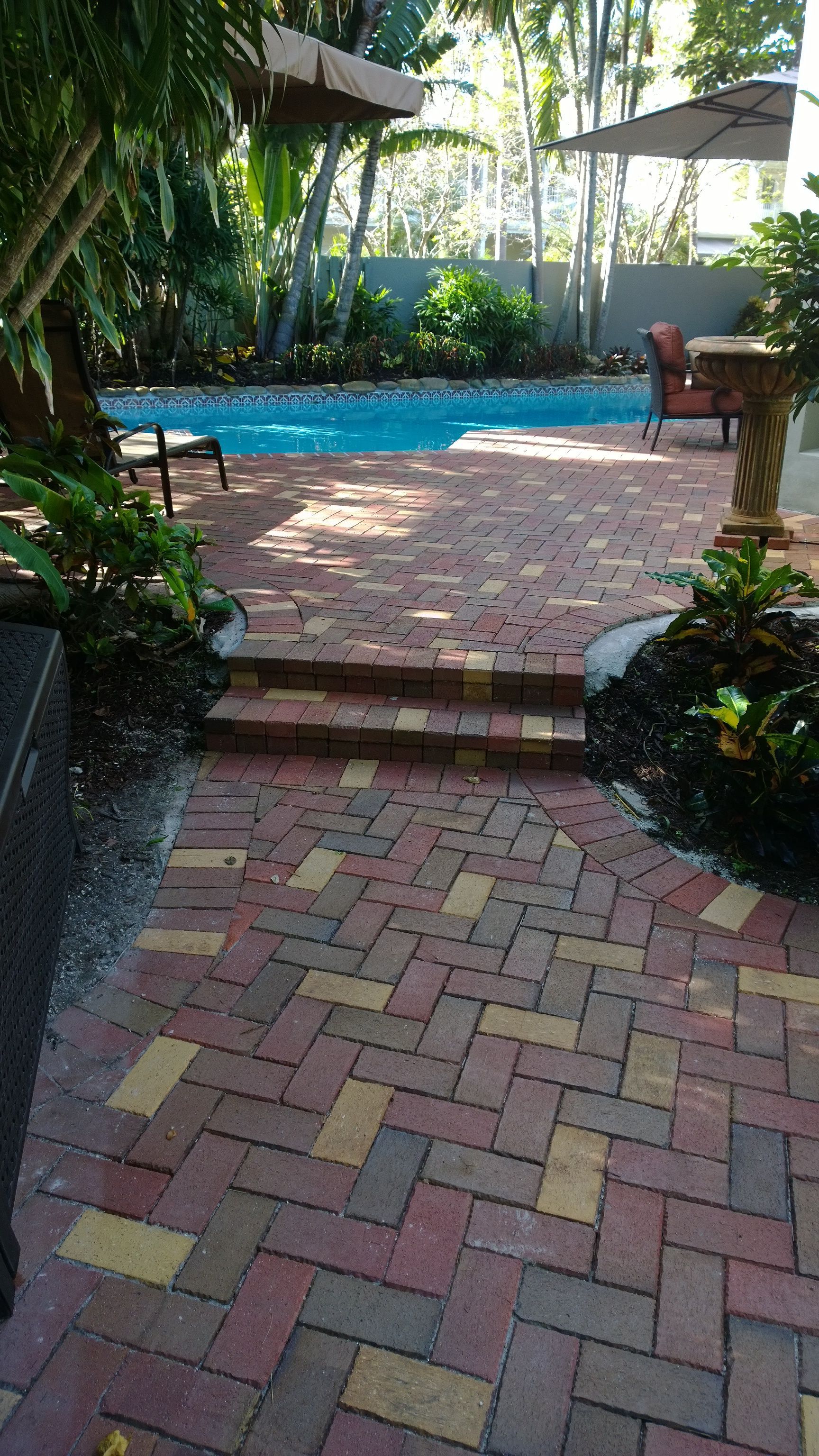 Brick patio path leading to a backyard pool, surrounded by lush greenery and shaded seating