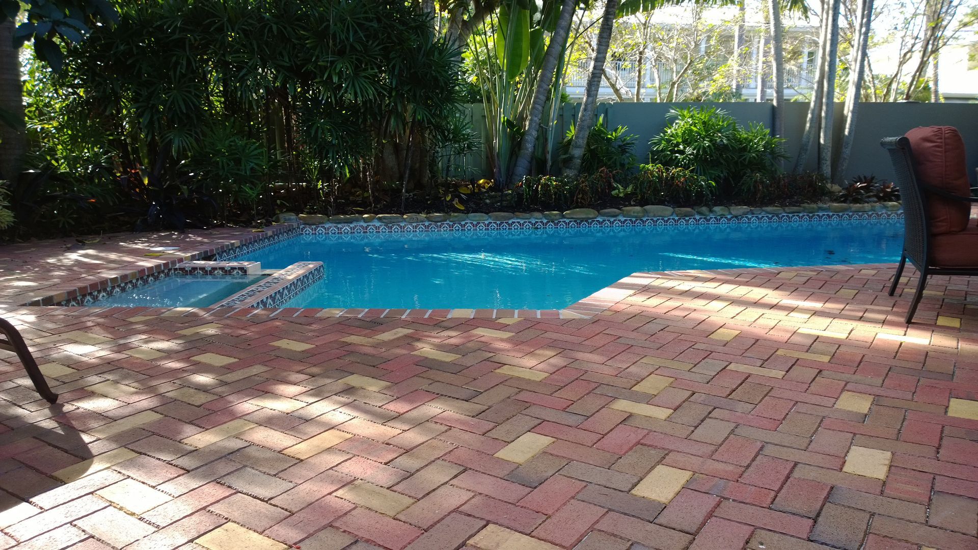Swimming pool next to a building, blue water, tiled patio, plants and blue sky.