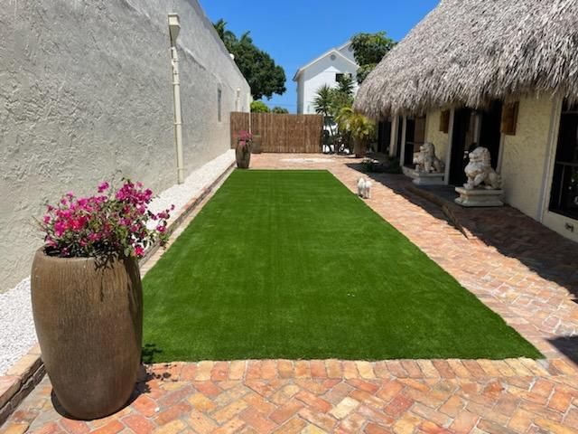Backyard patio with artificial grass, stone walkway, and potted flowers beside a thatched-roof house
