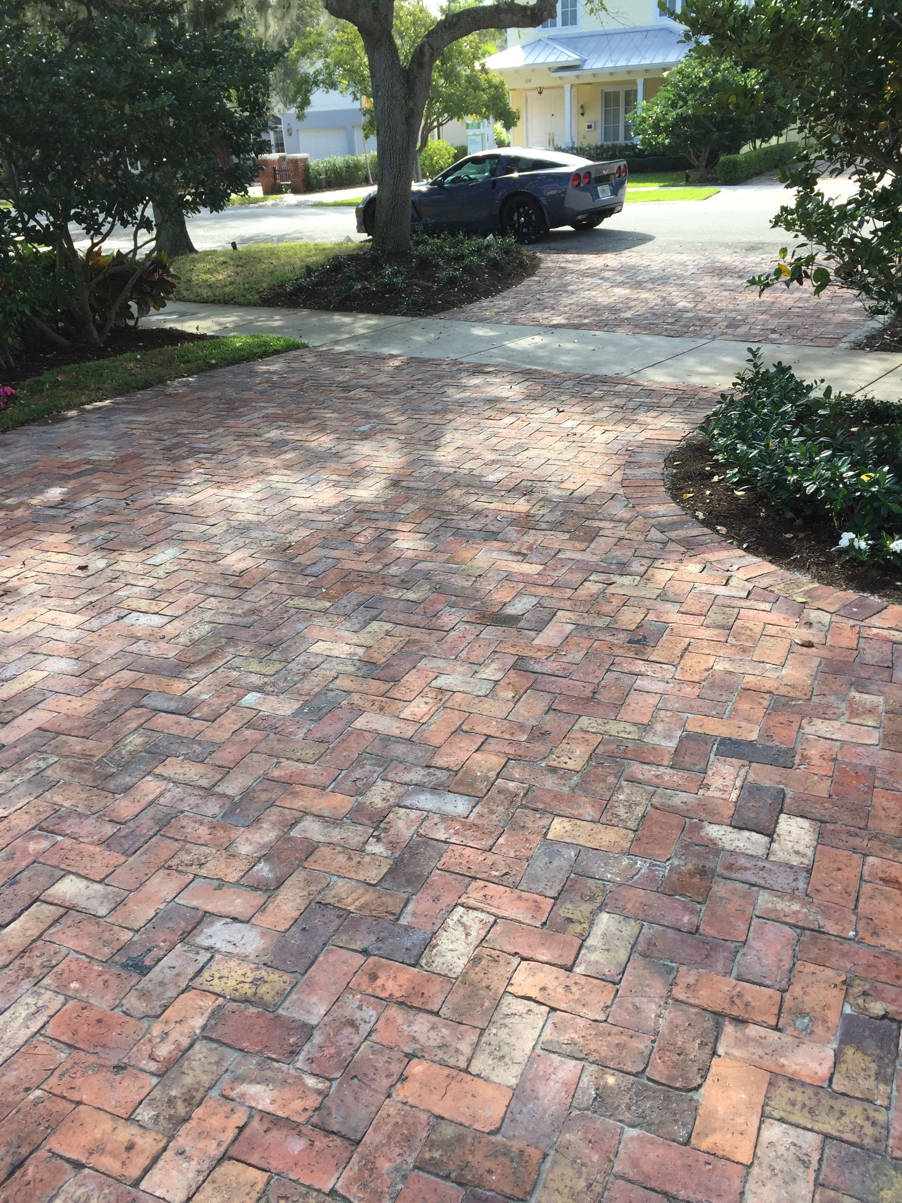Brick driveway leading to a house, with trees, shrubs, and a parked black car in the background