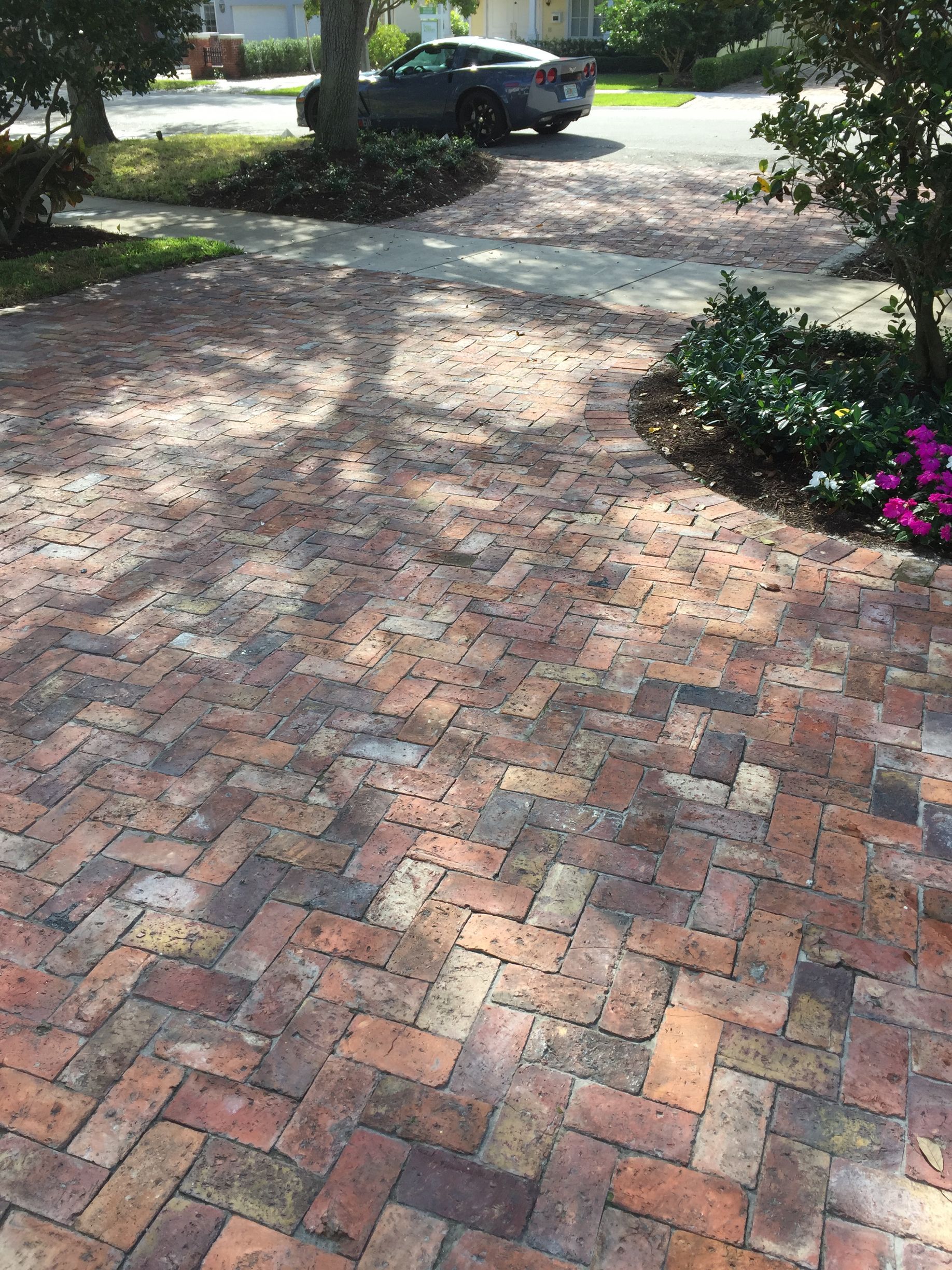 Brick driveway in dappled sunlight with a parked car and flower beds at the edge