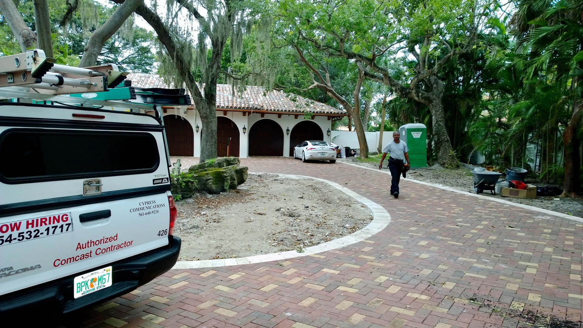 Tree-lined brick driveway leading to a building with arched porch, with a parked white van in the foreground.