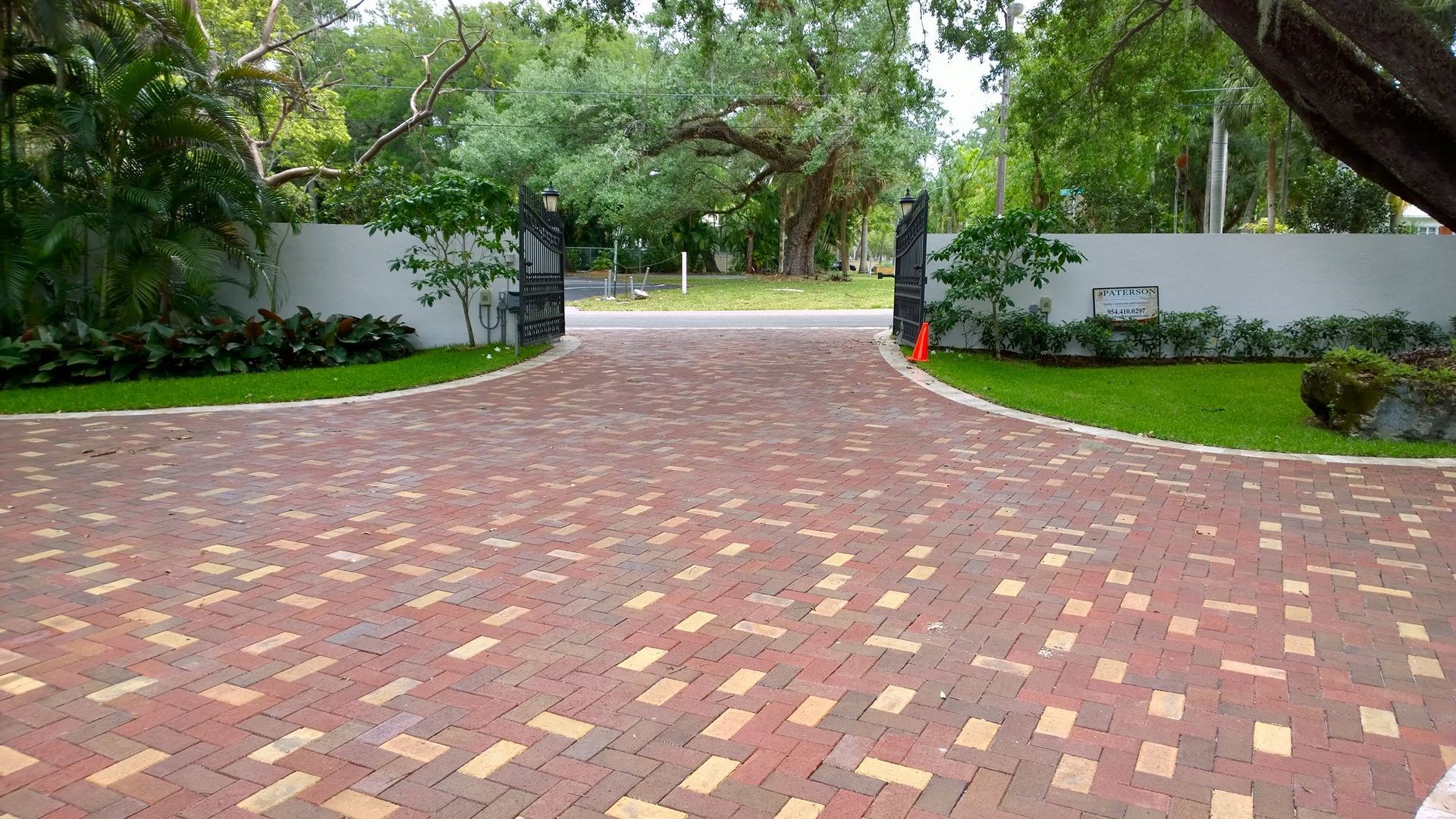 Brick-paved driveway leading to an open gated entrance surrounded by green trees and hedges