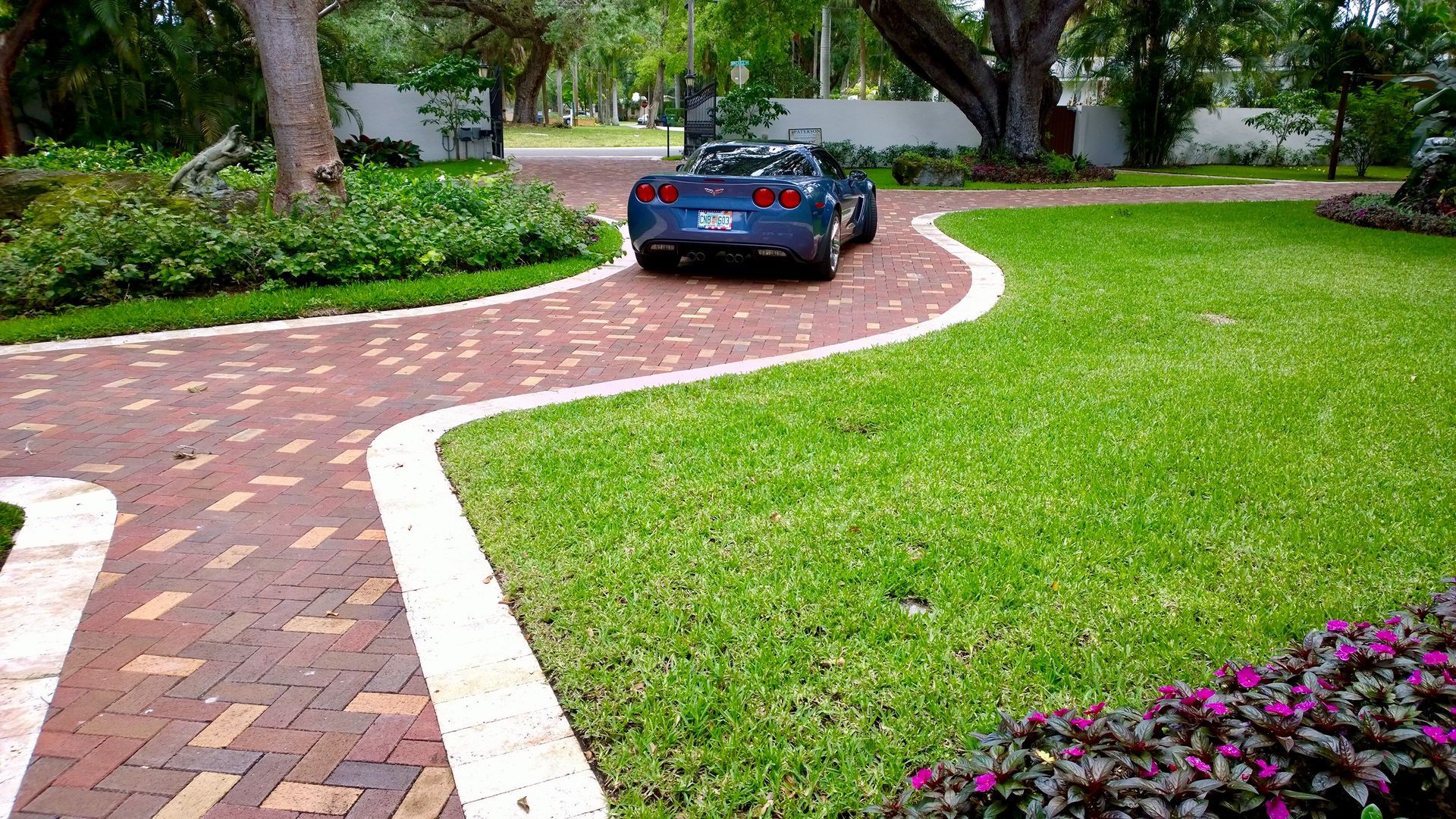 Blue sports car on a winding brick driveway through a landscaped garden with green lawn and trees.