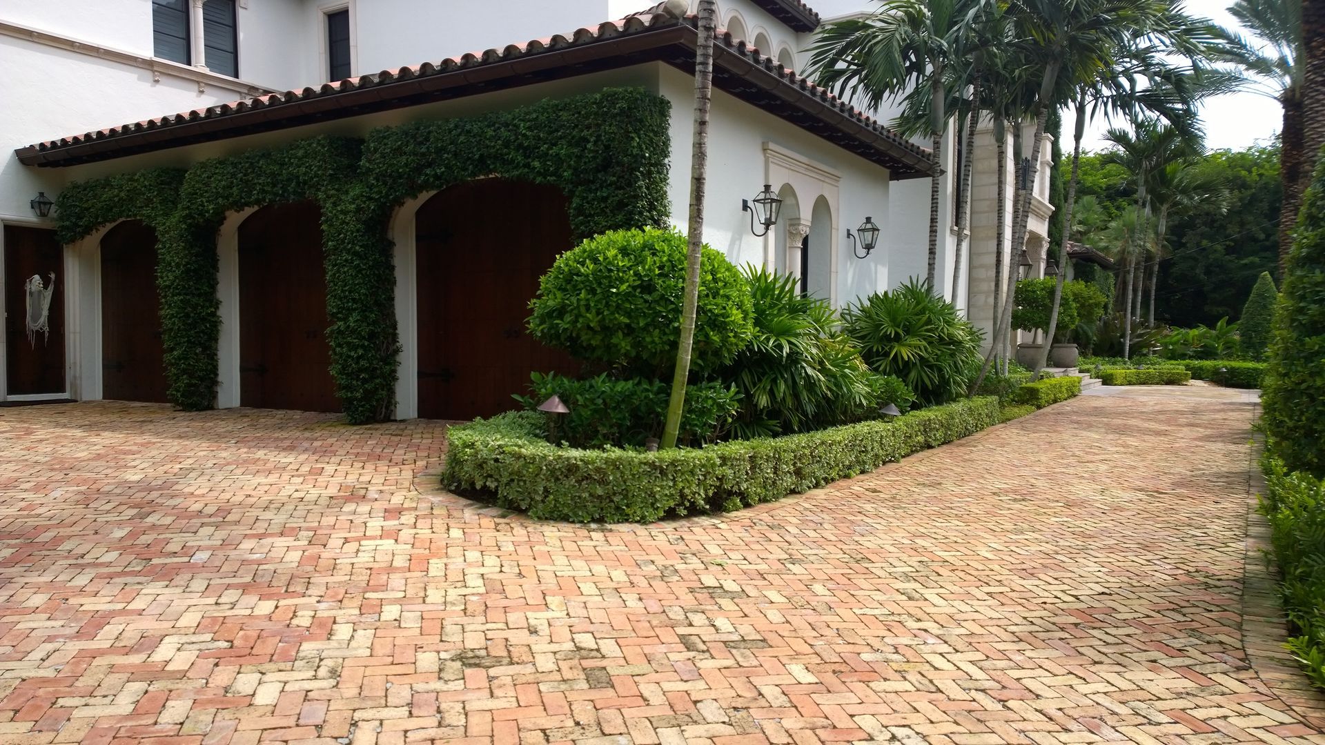 White villa with ivy, arched porch, and brick driveway surrounded by lush green landscaping