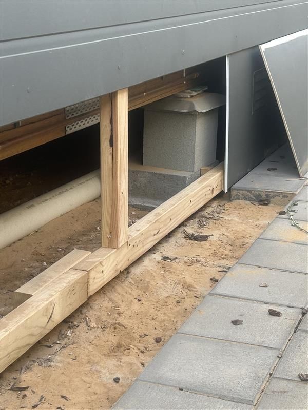 Open utility opening under a gray wall, braced with wooden supports and exposed pipes beside a sidewalk.