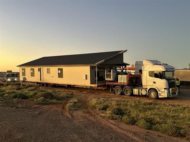 A white semi-truck hauling a long house on a rural road at sunset.