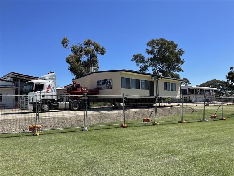 Mobile home being transported on a flatbed truck beside a fenced grassy lot under a clear blue sky Mobile home being transported on a flatbed truck beside a fenced grassy lot under a clear blue sky